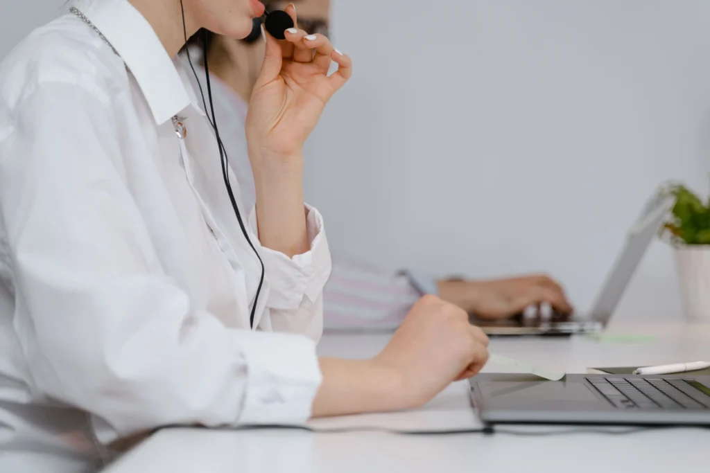 Sales development representative on a B2B telemarketing call using a headset, with a colleague working on a laptop beside her