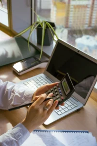Person using a calculator and pen at a desk with a laptop and notebook, working out the cost of a B2B telemarketing campaign